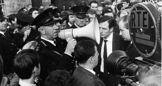 The photography is in black and white. A large crowd can be seen gathered around a group of five RUC policemen. An RTÉ camera can be seen in the foreground. County Inspector William Meharg is centre frame and uses a megaphone to warn the demonstrators in the crowd. This photo is from the Derry civil rights march on 5 October 1968 and is considered to be the start of the Troubles. Credit: The Irish Times
